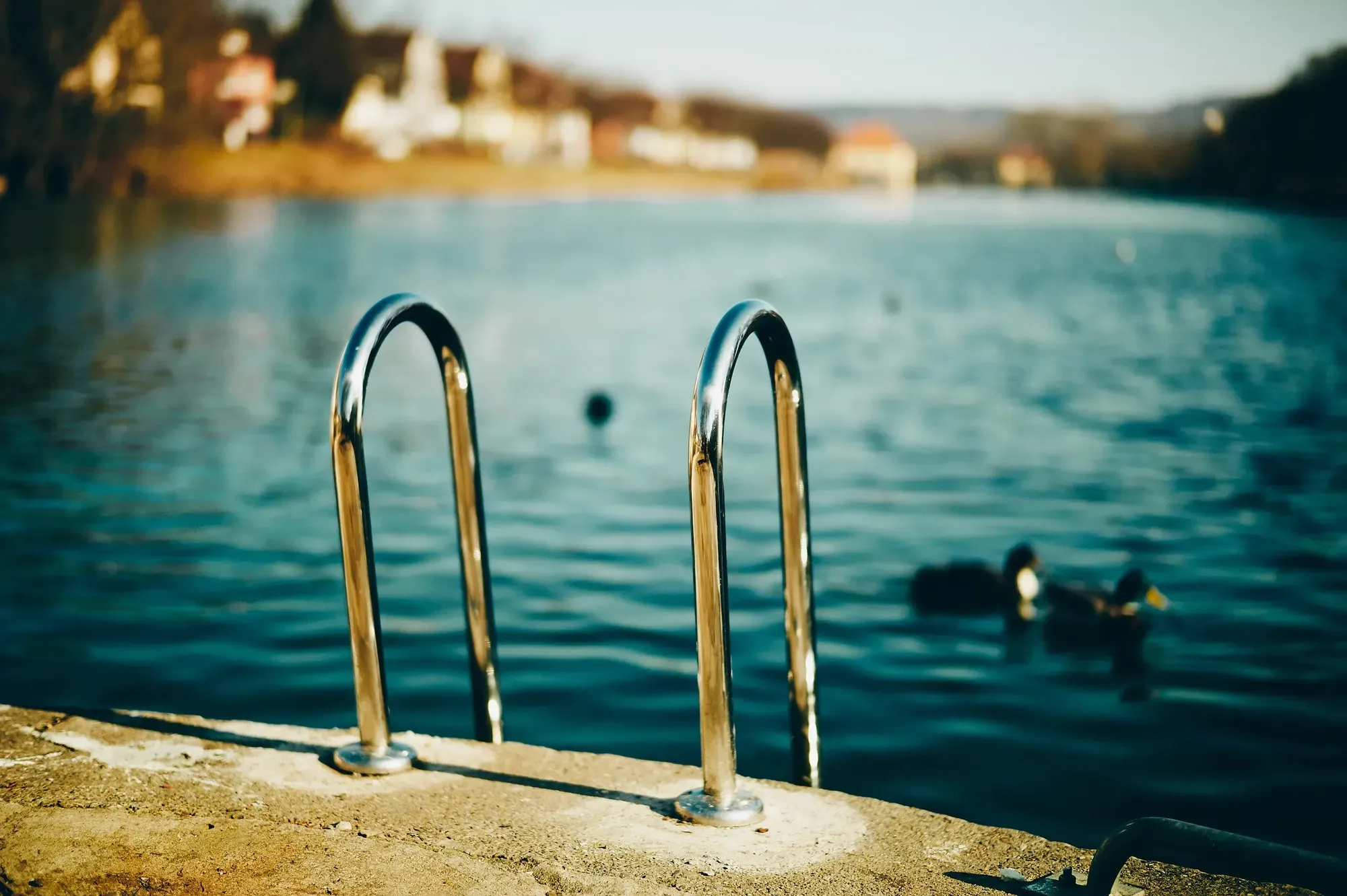  Auf dem Bild ist alles andere als das zu sehen, was üblicherweise unter einem Hafen erwartet werden darf. Tübingens Anlegestellen für Stocherkähne liegen unter anderem an der Mündung der Steinlach in den Neckar. Für den Fall, dass Menschen ins Wasser fallen oder ein Stocherkahn umständlich beladen werden müsste, gibt es im Steinlachhafen eine Trittleiter aus chromiertem Stahl die vom betonierten, bräunlich schimmernden Steg aus zum blaugrünlich wirkenden Wasser des Neckars herunterreicht. Diese Trittleiter ist im Zentrum der Aufnahme zu sehen. Der Chromeüberzug blitzt ein wenig im Sonnenlicht. Im Hintergrund ist die Uferböschung mit dahinterliegenden Häusern unscharf zu erkennen, wobei sich auf dem Neckar ein paar Stockenten treiben lassen.