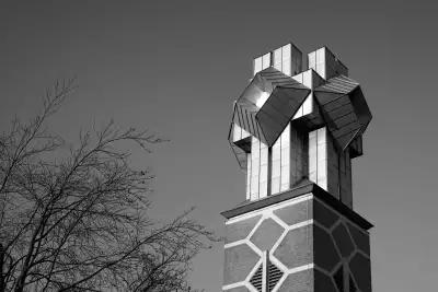 Schwarz-Weiß Aufnahme. Zeigt eine neuzeitlich gestaltete und glänzende Kirschturmspitze in Form eines Kreuzes mit aufgesetzen Quadraten, in deren Oberfläche sich die Sonnenstrahlen spiegeln.