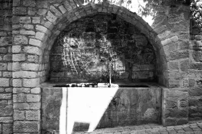 Schwarz-Weiß Aufnahme; zeigt einen alten, von der Sonne angestrahlten Quellbrunnen am Straßenrand.