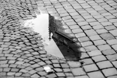 Schwarz-Weiß Aufnahme; zeigt eine Regenpfütze auf Kopfsteinpflaster in welches sich ein Teil der Dachkonstruktion der "Holzmarktkirche" aus Tübingen spiegelt.