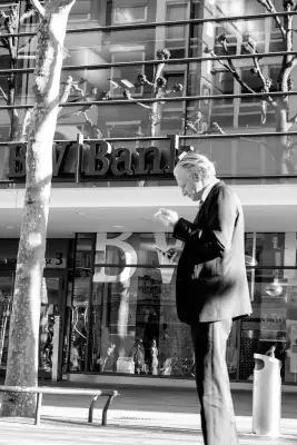 Schwarz-Weiß Aufnahme; Zeigt einen Mann mit Zigarette in der Hand und dem Blick auf das Smartphone gerichtet, der in einer Fußgängerzone vor der "BW-Bank" steht.