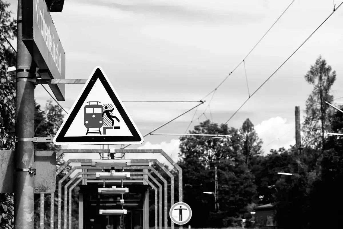 Schwarz-Weiß Aufnahme; zeigt ein Warnschild vor herannahenden Zügen bzw. der Sturzgefahr an einer Bahnsteigkante.