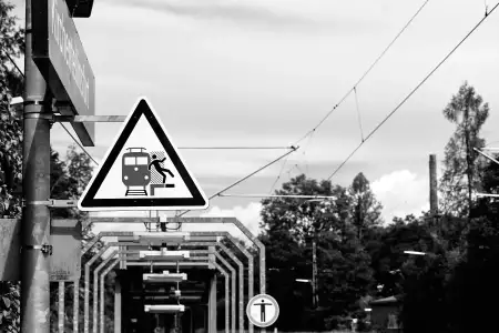 Schwarz-Weiß Aufnahme; zeigt ein Warnschild vor herannahenden Zügen bzw. der Sturzgefahr an einer Bahnsteigkante.