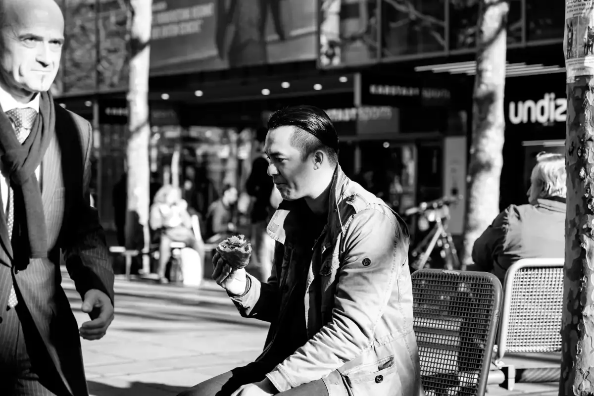 Schwarz-Weiß Aufnahme; zeigt einen jungen Mann in Freizeitkleidung und ein Brötchen essend auf einer Bank sitzend und einen älteren Mann im 