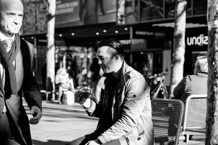 Schwarz-Weiß Aufnahme; zeigt einen jungen Mann in Freizeitkleidung und ein Brötchen essend auf einer Bank sitzend und einen älteren Mann im 