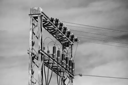 Schwarz-Weiß Aufnahme; zeigt die an einer Verteilerstation beginnenden, zahlreichen Stromleitungen und ihre Isolatoren, angebracht an einem bereits rostenden Metallträger.