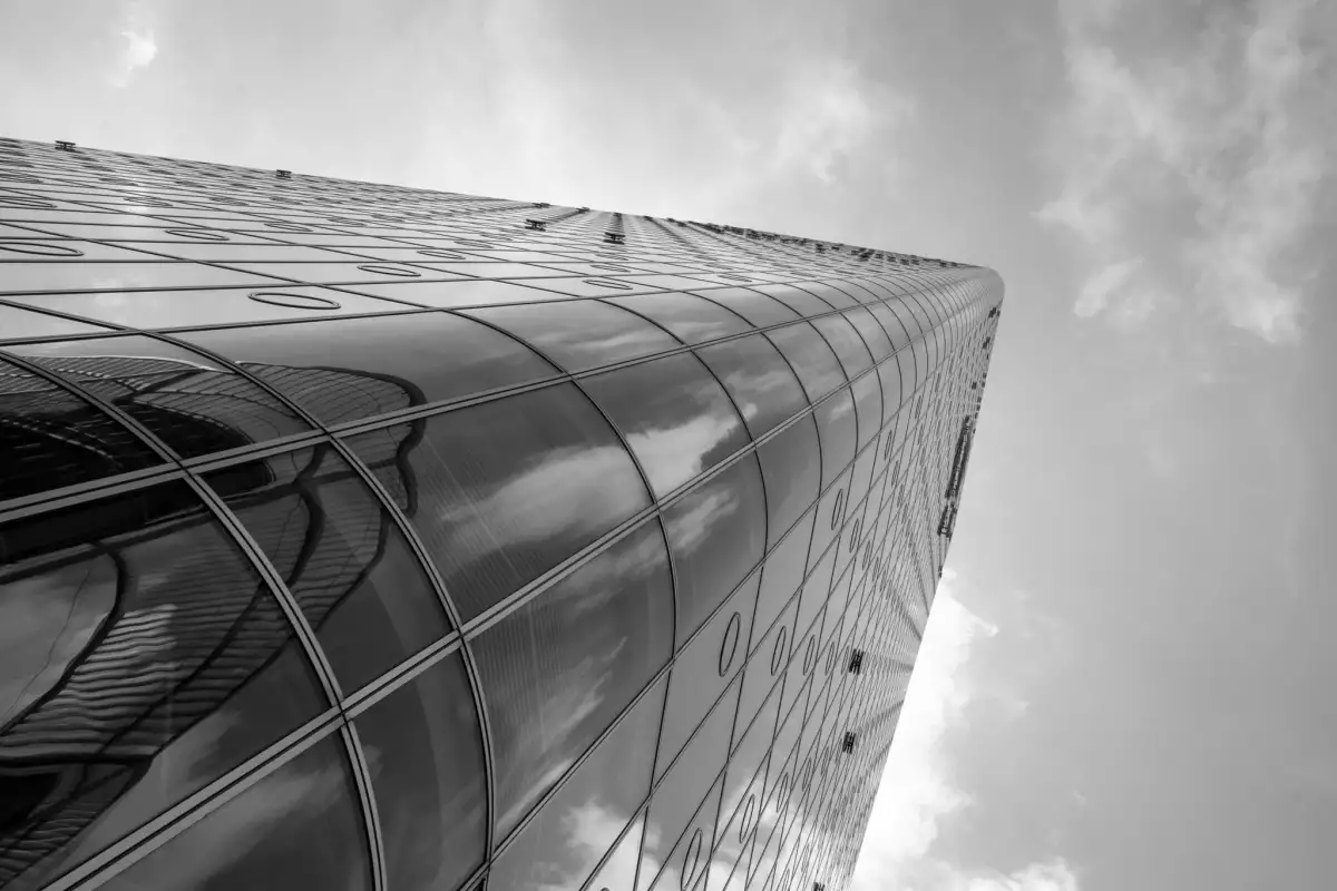Schwarz-Weiß Aufnahme. Zeigt von unten nach oben gerichtet die Glasfassade eines Bürohochhauses in München auf der sich die Wolken spiegeln.