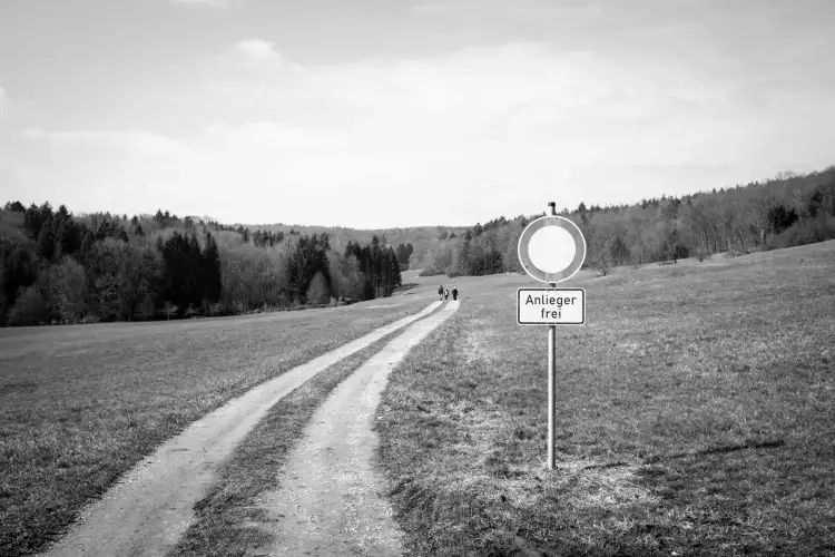 Schwarz-Weiß Aufnahme; zeigt ein Feldweg, der entlang einer Wiese in ein Waldgebiet führt, wobei am Anfang ein Straßenschild zu lesen ist, auf dem steht: 